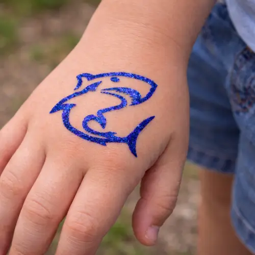 Tatouage pailleté requin : Un tatouage éphémère réalisé avec un pochoir adhésif, de la colle cosmétique et des paillettes.