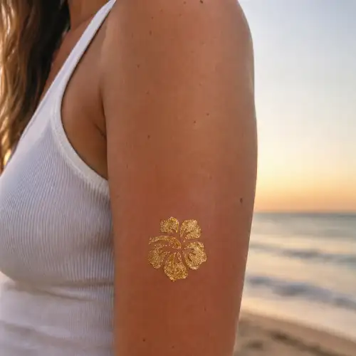 Tatouage paillette hibiscus doré appliqué sur le haut du bras, réalisé à l'aide d'un pochoir et de colle cosmétique.