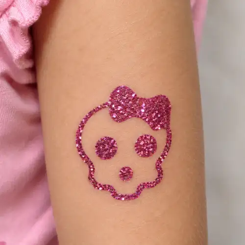 Tatouage éphémère pailleté rose représentant une tête de mort girly avec un nœud. Le motif est réalisé au pochoir avec de la colle cosmétique et des paillettes brillantes.